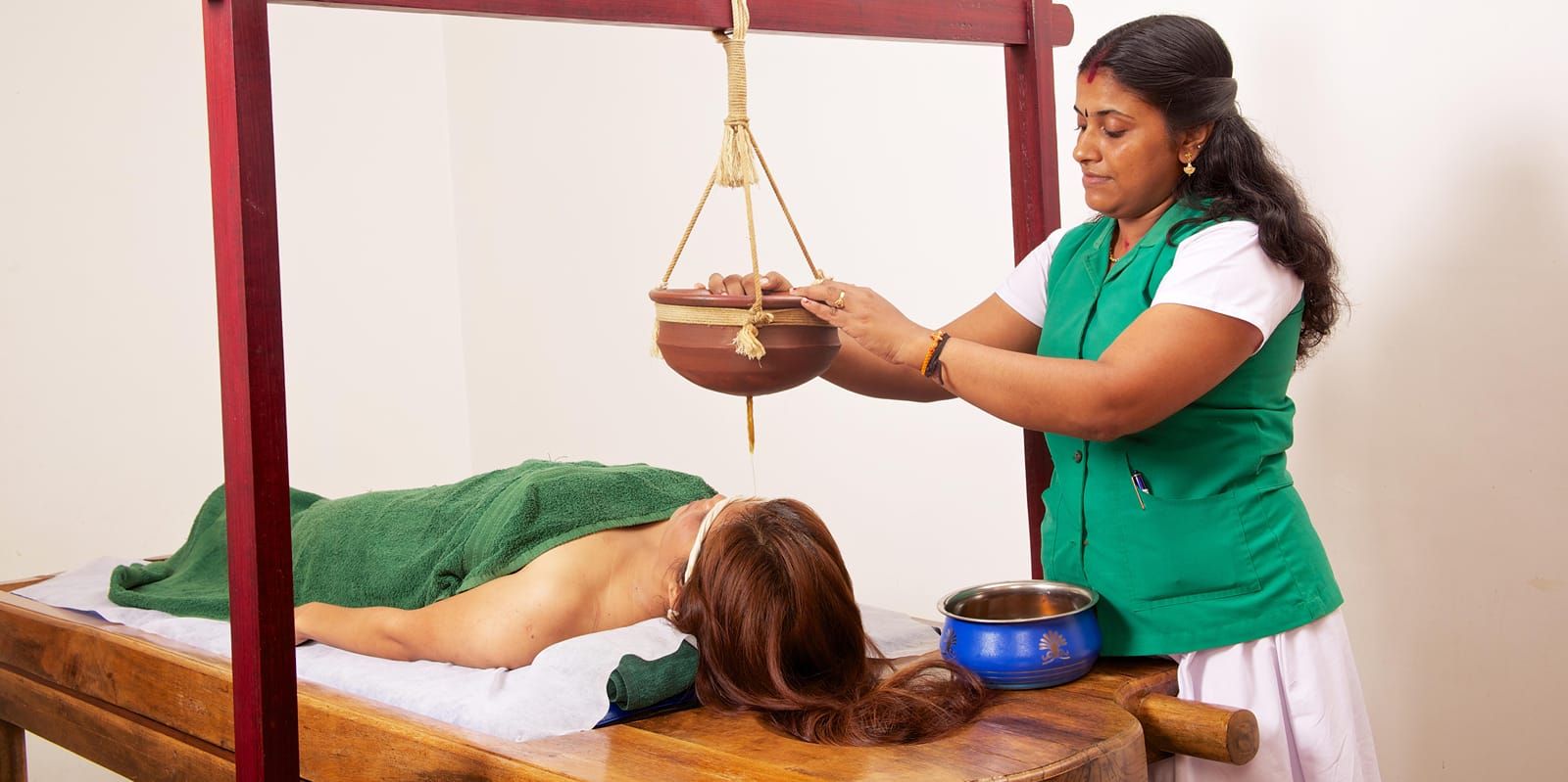 A therapist adjusting a hanging wooden pot over a person's forehead during a traditional Ayurvedic oil treatment